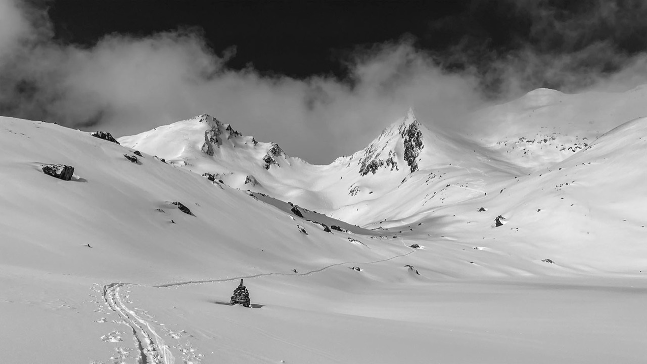 Snow-covered mountain landscape with a marked trail, a small pile of stones in the foreground, and dark clouds over the peaks.