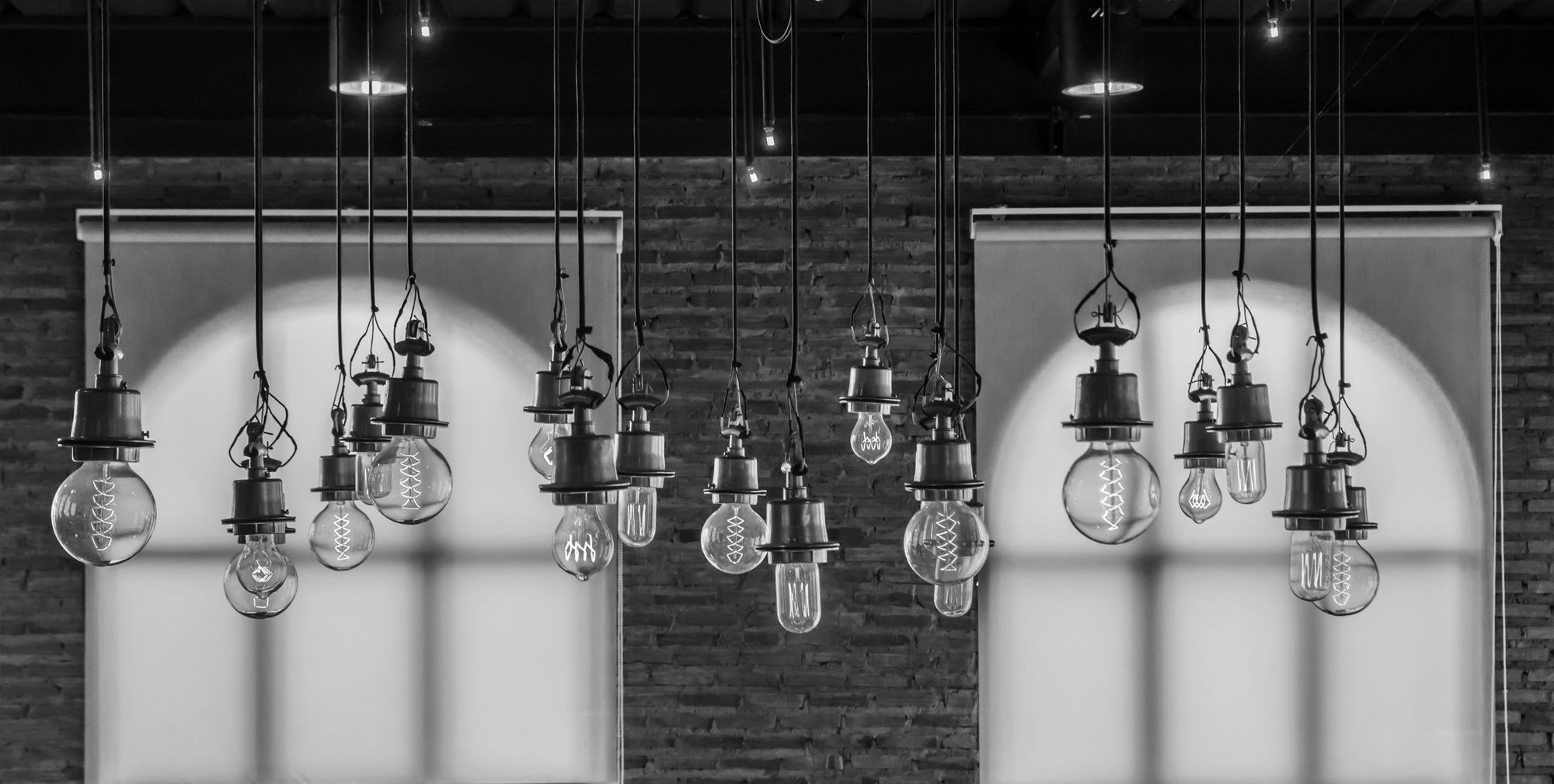 Black-and-white image with numerous hanging light bulbs in front of a brick wall and two bright wall sections.