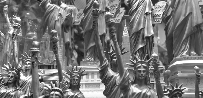 Black-and-white image featuring numerous miniature and large depictions of the Statue of Liberty against a cityscape of skyscrapers.