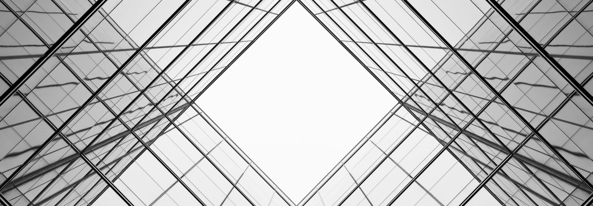 Black-and-white image of a glass building facade with intersecting steel beams and a bright, diamond-shaped patch of sky in the center.
