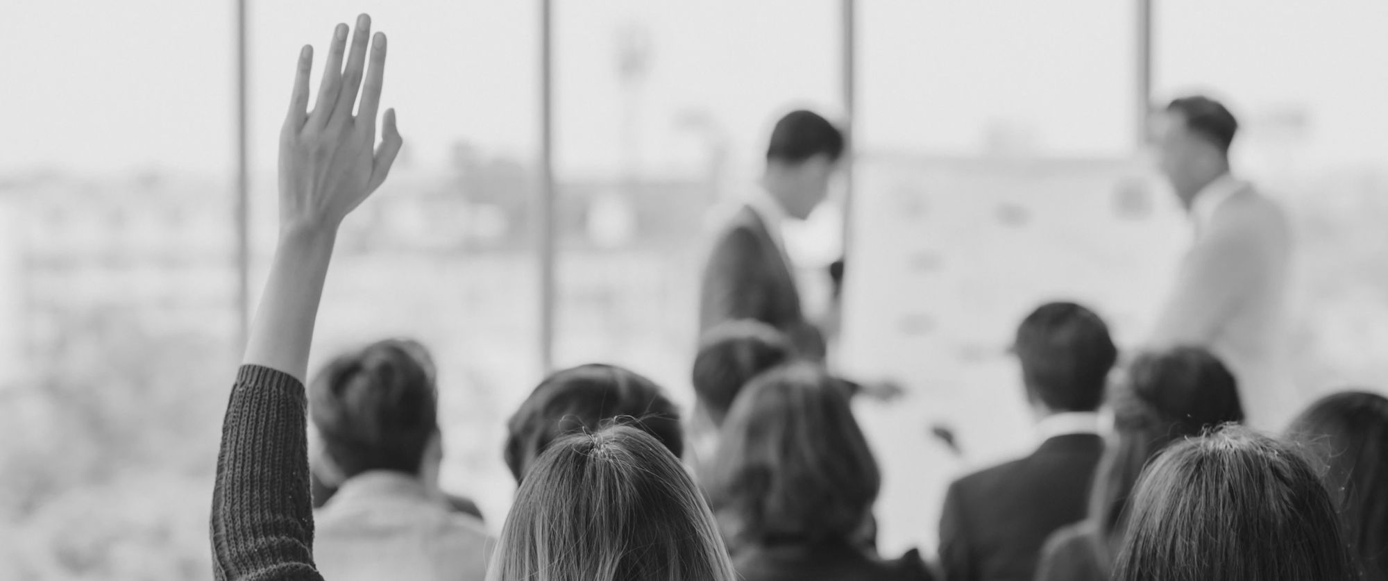 Black-and-white photo of a group of people in a seminar room; one person raises a hand in the foreground.