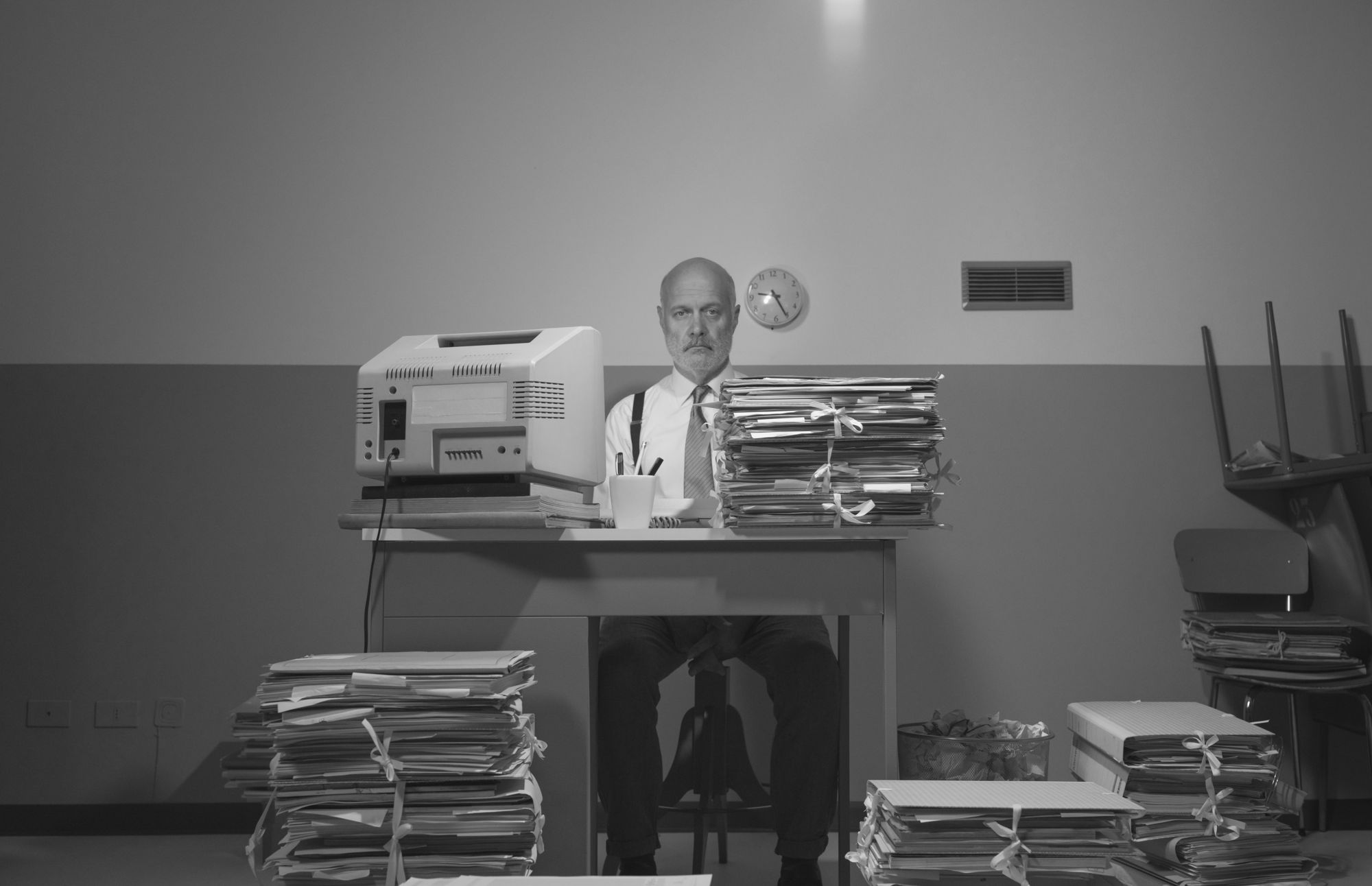 Black-and-white image of a serious-looking office worker behind a desk, surrounded by tall stacks of bound files and an old computer monitor.