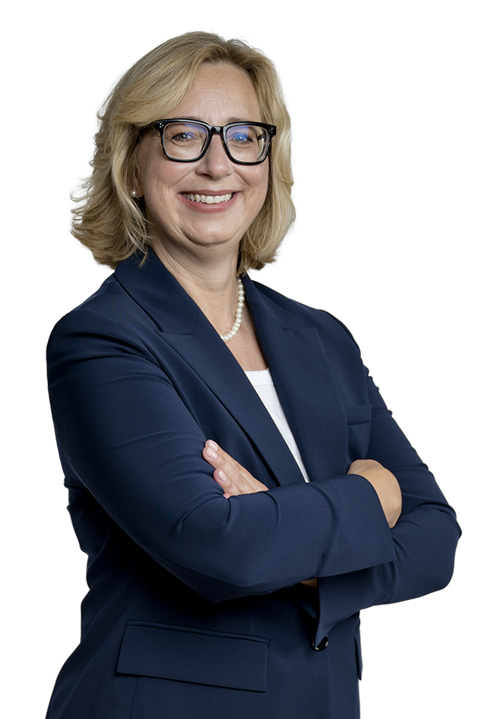 Porträt einer lächelnden blonden Frau mit Brille, Perlenkette und dunkelblauem Blazer, die verschränkt die Arme hält.
