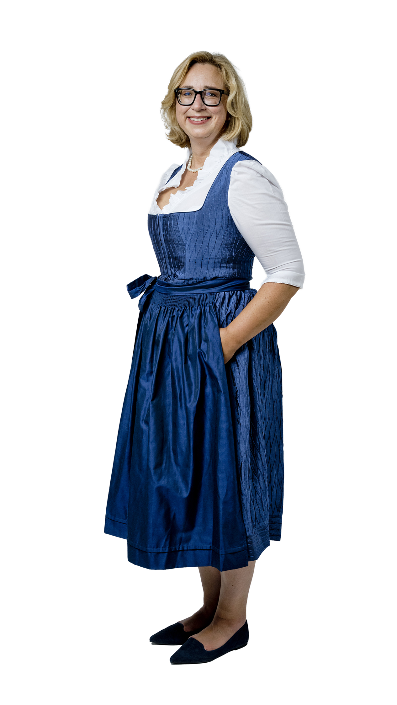 Eine lächelnde Frau mit Brille und blonden Haaren trägt ein blaues Dirndl mit weißer Bluse und steht vor hellem Hintergrund.
