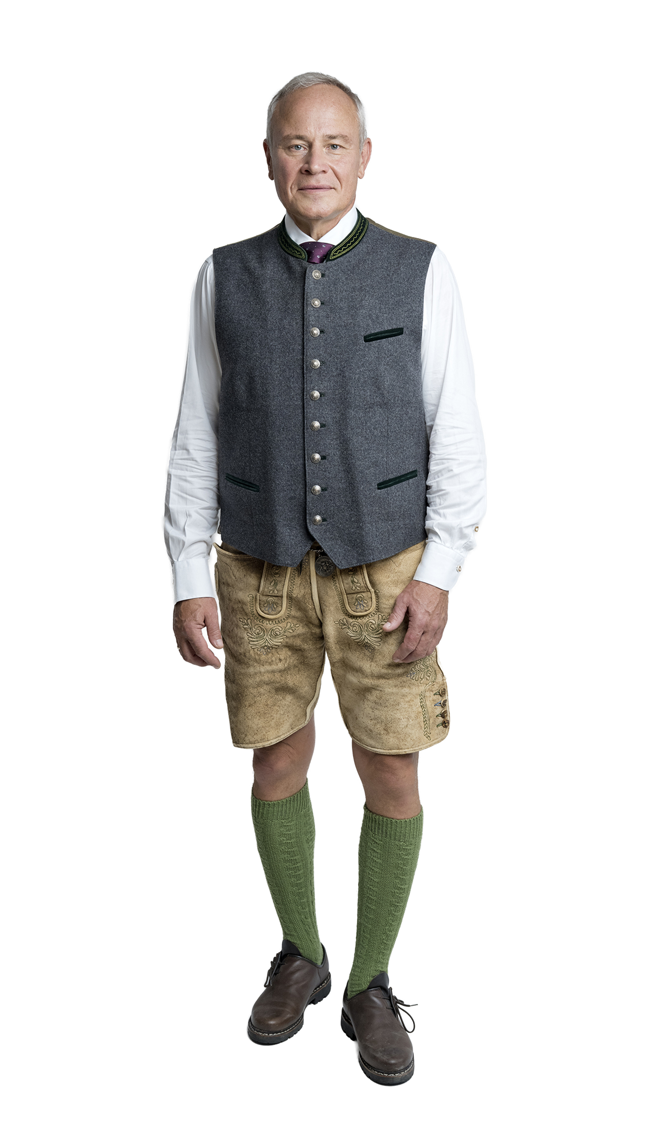 Ganzkörperaufnahme eines Mannes in traditioneller bayerischer Tracht mit grauer Weste, weißem Hemd, bestickten Lederhosen, grünen Kniestrümpfen und braunen Haferlschuhen.