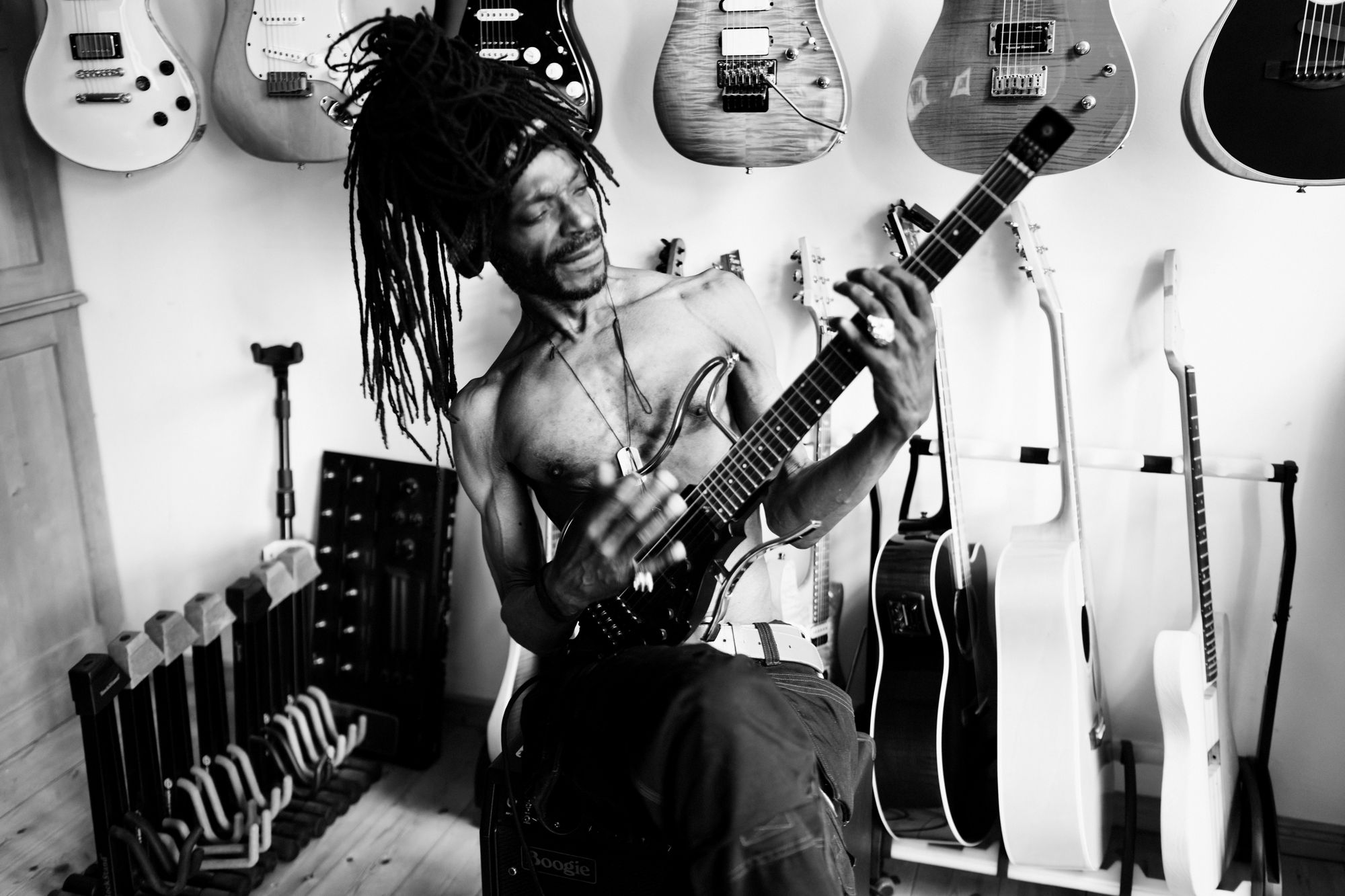 A man with long dreadlocks plays an electric guitar in a room surrounded by guitars.