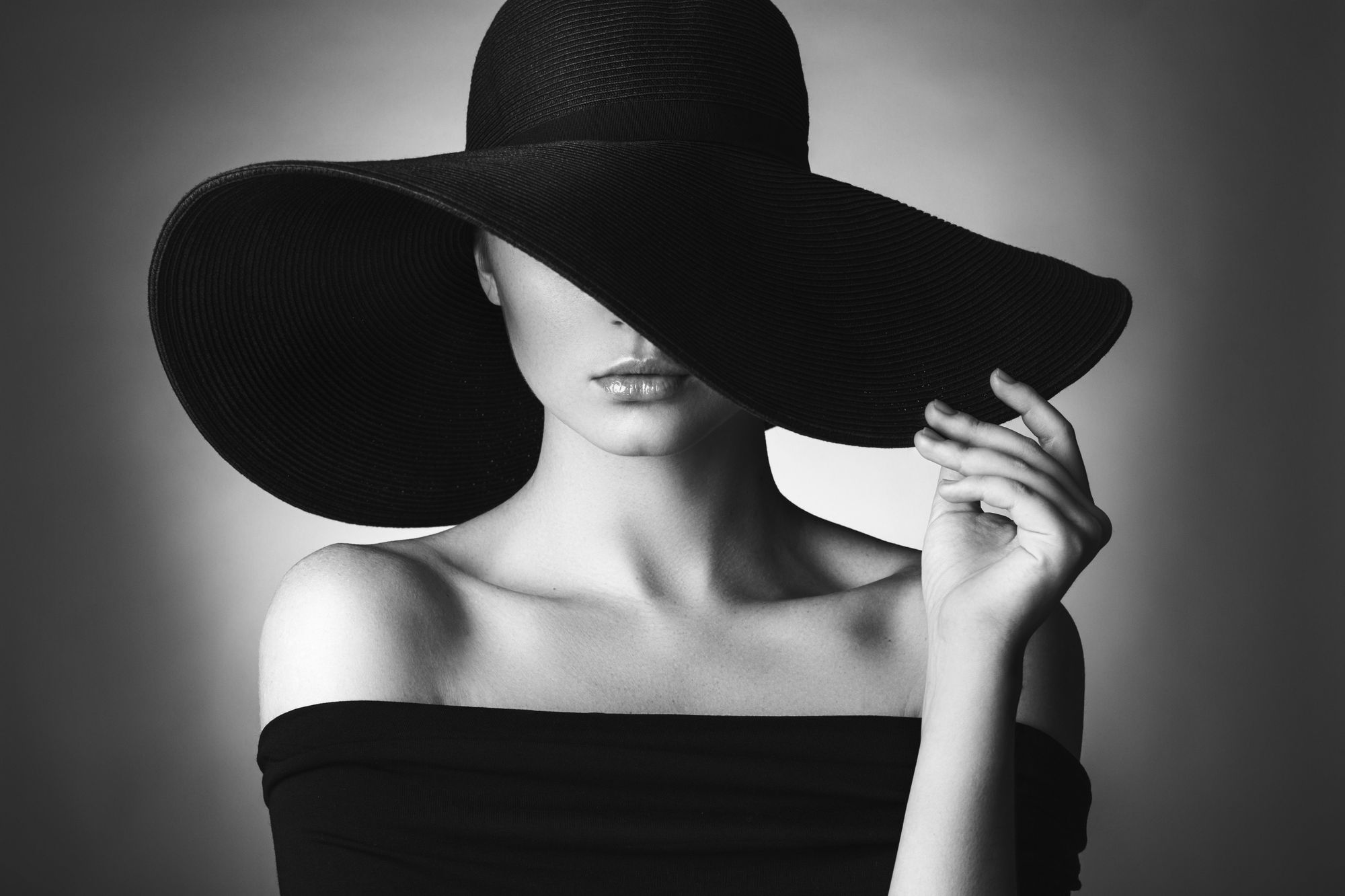 Black-and-white portrait of a woman wearing a large black hat with a wide brim partially covering her eyes; she wears an off-the-shoulder top and touches the brim.