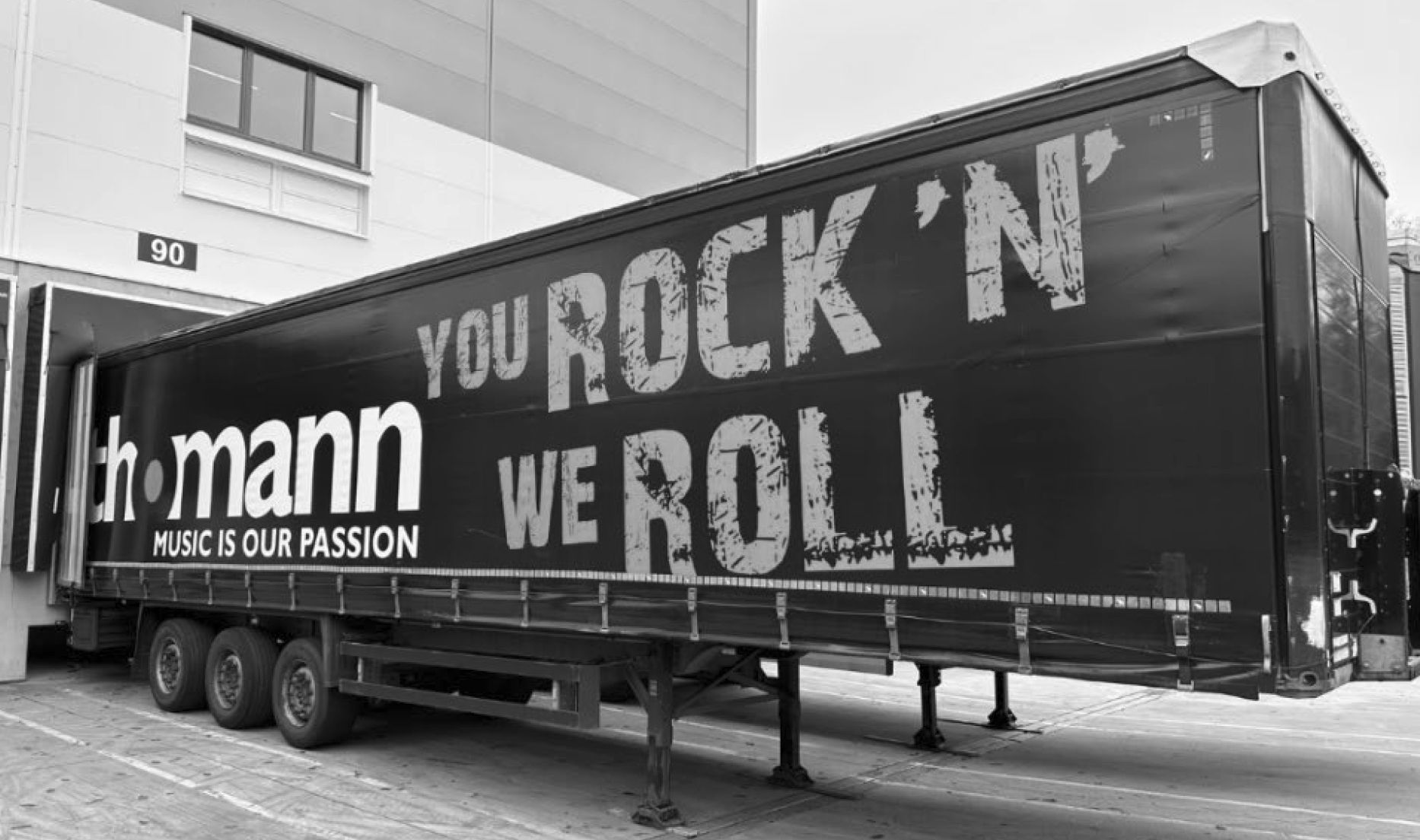 Black-and-white image of a large semi-trailer bearing the words “YOU ROCK ’N’ ROLL” and “Music Is Our Passion” in front of a building.