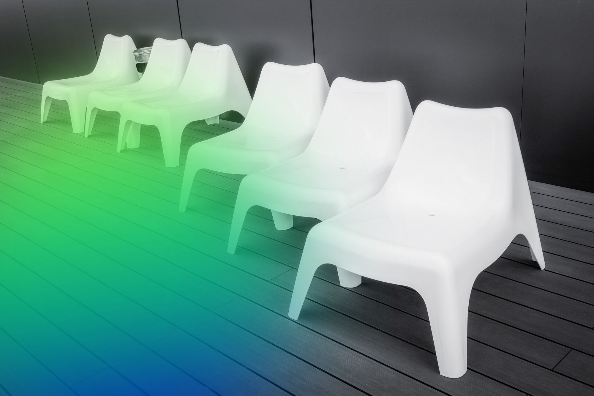 Several white plastic loungers stand on a dark wooden terrace in front of a black wall; green and blue light illuminates the floor.